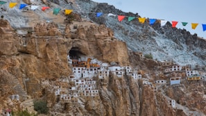 A series of buildings are nestled into a rocky cliffside, blending with the natural rock formations. These structures appear to be traditional, with some elements painted in white and other vibrant colors. Above, strings of colorful prayer flags flutter in the wind, adding a vibrant touch against the rugged terrain.