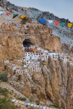 A cluster of white buildings with red roofs is nestled into a rocky cliffside, creating a dramatic architectural feature. Colorful prayer flags are strung across the sky, adding vibrant tones above the earthy rock formations. The rugged terrain surrounds the unique settlement, blending natural beauty with human ingenuity.