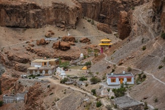 A remote hillside settlement features traditional buildings with flat roofs and colorful trims, surrounded by rugged, rocky terrain. The structures include a small temple with yellow and red accents perched against the barren landscape. Sparse vegetation dots the rocky slopes, and a few pathways connect the isolated structures.