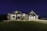 A large modern house illuminated at night, featuring a combination of stone and siding exterior. It has multiple peaked roofs, a spacious driveway leading to a three-car garage, and well-maintained landscaping surrounding the front.