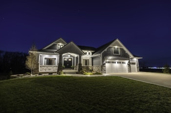 A large modern house illuminated at night, featuring a combination of stone and siding exterior. It has multiple peaked roofs, a spacious driveway leading to a three-car garage, and well-maintained landscaping surrounding the front.