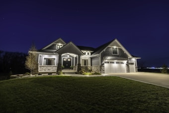 A large modern house illuminated at night, featuring a combination of stone and siding exterior. It has multiple peaked roofs, a spacious driveway leading to a three-car garage, and well-maintained landscaping surrounding the front.