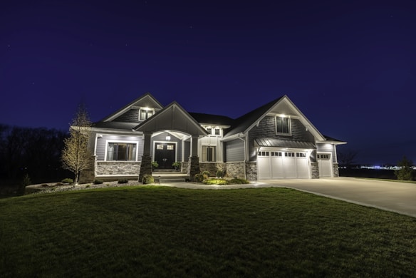 A large modern house illuminated at night, featuring a combination of stone and siding exterior. It has multiple peaked roofs, a spacious driveway leading to a three-car garage, and well-maintained landscaping surrounding the front.