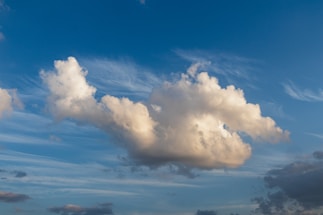 A large fluffy cloud dominates the center against a background of clear blue sky, with wisps of white clouds surrounding it. The cloud casts a soft, warm glow as it reflects sunlight.