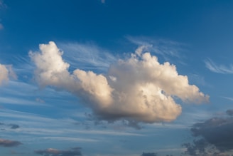 A large fluffy cloud dominates the center against a background of clear blue sky, with wisps of white clouds surrounding it. The cloud casts a soft, warm glow as it reflects sunlight.