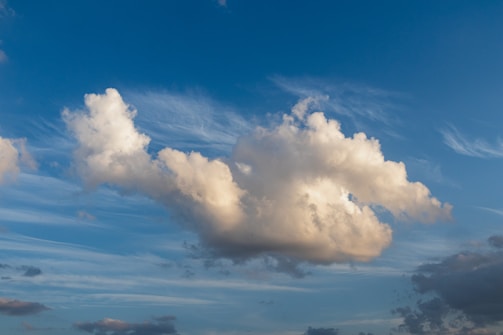 A large fluffy cloud dominates the center against a background of clear blue sky, with wisps of white clouds surrounding it. The cloud casts a soft, warm glow as it reflects sunlight.