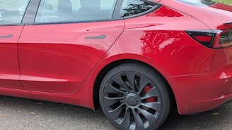 The image depicts the rear side of a red sedan vehicle, focusing on the back door, rear tire, and part of the back window. The car has sleek design lines, and the tire features a modern rim design.