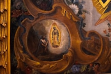 A detailed painting depicts a religious scene with a central figure illuminated by an intricate golden frame. Surrounding the figure are rich floral decorations and swirling ornate patterns in warm tones. The artwork conveys a sense of devotion and reverence.