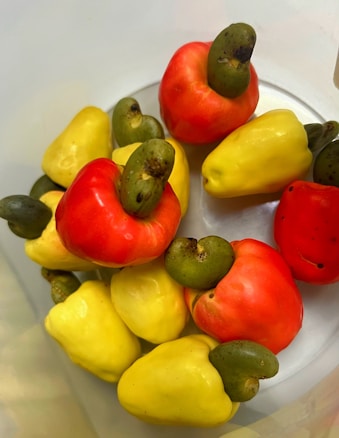 A collection of ripe and colorful cashew fruits, with a combination of yellow and red cashew apples, each topped with a green cashew nut. The fruits are placed together in a light-colored container, creating a vibrant contrast.