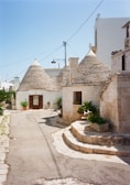 A picturesque street featuring traditional stone houses with conical roofs, known as trulli. The stone buildings are complemented by green plants in pots, adding a touch of nature. The pathway is narrow and paved with stones, leading through this charming architectural setting.