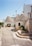 A picturesque street featuring traditional stone houses with conical roofs, known as trulli. The stone buildings are complemented by green plants in pots, adding a touch of nature. The pathway is narrow and paved with stones, leading through this charming architectural setting.