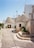 A picturesque street featuring traditional stone houses with conical roofs, known as trulli. The stone buildings are complemented by green plants in pots, adding a touch of nature. The pathway is narrow and paved with stones, leading through this charming architectural setting.