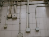 An interior wall is covered with a grid of beige tiles. Several electrical conduits run vertically and horizontally across the surface, connecting multiple junction boxes affixed to the wall. One of the boxes is marked with the letter 'N'. The overall appearance is industrial and functional.