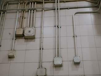 An interior wall is covered with a grid of beige tiles. Several electrical conduits run vertically and horizontally across the surface, connecting multiple junction boxes affixed to the wall. One of the boxes is marked with the letter 'N'. The overall appearance is industrial and functional.