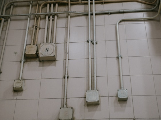 An interior wall is covered with a grid of beige tiles. Several electrical conduits run vertically and horizontally across the surface, connecting multiple junction boxes affixed to the wall. One of the boxes is marked with the letter 'N'. The overall appearance is industrial and functional.