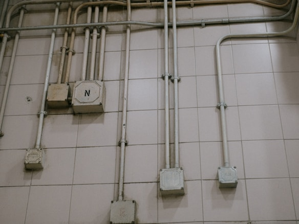 An interior wall is covered with a grid of beige tiles. Several electrical conduits run vertically and horizontally across the surface, connecting multiple junction boxes affixed to the wall. One of the boxes is marked with the letter 'N'. The overall appearance is industrial and functional.