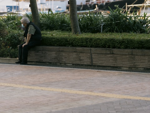 An elderly person wearing a mask sits alone on a low brick wall surrounded by a neatly trimmed hedge and potted plants. The background shows a blurred urban scene with trees and a waterfront area.