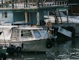 A person stands on a small boat docked near a larger structure on the water. The individual is holding two plastic bottles and appears to be shirtless, wearing dark shorts and a white cap. The scene includes multiple boats, some with canopies, in a marina-like setting. The water reflects light, adding a shimmering effect, and the structures show signs of wear.