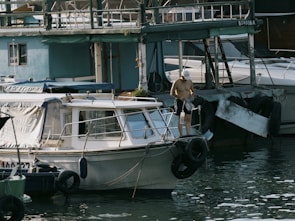 A person stands on a small boat docked near a larger structure on the water. The individual is holding two plastic bottles and appears to be shirtless, wearing dark shorts and a white cap. The scene includes multiple boats, some with canopies, in a marina-like setting. The water reflects light, adding a shimmering effect, and the structures show signs of wear.