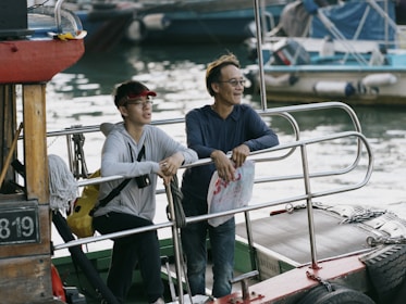 Two people are standing on a boat, leaning on the railing and facing towards the water. They are engaged in a relaxed conversation. One person is wearing a red visor, white zip-up jacket, and has a yellow backpack slung over their shoulder. The other person is wearing glasses and a blue jacket, holding a plastic bag. The surrounding area includes other boats and water, indicating a dock or marina setting.