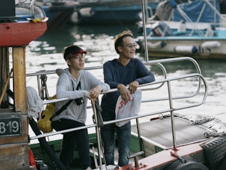 Two people are standing on a boat, leaning on the railing and facing towards the water. They are engaged in a relaxed conversation. One person is wearing a red visor, white zip-up jacket, and has a yellow backpack slung over their shoulder. The other person is wearing glasses and a blue jacket, holding a plastic bag. The surrounding area includes other boats and water, indicating a dock or marina setting.