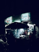 A dimly lit street scene at night featuring a medical store with a glowing sign. Several people stand or walk near the entrance, while a street food vendor with an umbrella is visible in the foreground. The setting is urban, with a few motorbikes parked on the side.