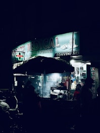 A dimly lit street scene at night featuring a medical store with a glowing sign. Several people stand or walk near the entrance, while a street food vendor with an umbrella is visible in the foreground. The setting is urban, with a few motorbikes parked on the side.