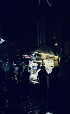 A night market scene with several silhouetted figures standing around a brightly lit vendor stall. The stall is selling perfumes and has numerous small bottles displayed. The ground appears wet, reflecting some of the lighting from the stall.