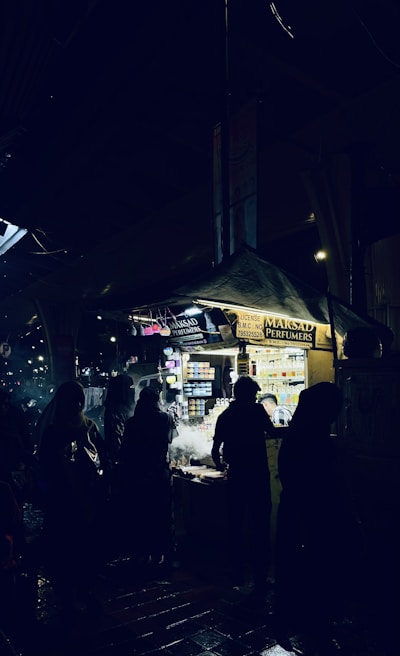 A night market scene with several silhouetted figures standing around a brightly lit vendor stall. The stall is selling perfumes and has numerous small bottles displayed. The ground appears wet, reflecting some of the lighting from the stall.