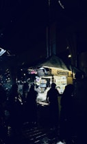 A night market scene with several silhouetted figures standing around a brightly lit vendor stall. The stall is selling perfumes and has numerous small bottles displayed. The ground appears wet, reflecting some of the lighting from the stall.