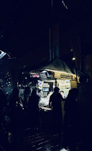 A night market scene with several silhouetted figures standing around a brightly lit vendor stall. The stall is selling perfumes and has numerous small bottles displayed. The ground appears wet, reflecting some of the lighting from the stall.