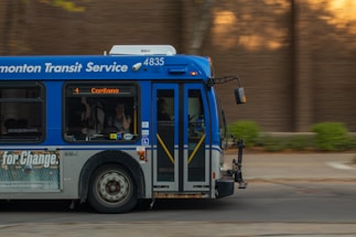 A public transit bus from the Edmonton Transit Service is captured in motion. The side of the bus shows advertisements and an electronic display indicates the destination as Capilano with route number 4. Inside, passengers are visible through the windows as they travel.
