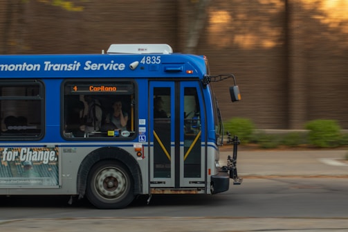 A public transit bus from the Edmonton Transit Service is captured in motion. The side of the bus shows advertisements and an electronic display indicates the destination as Capilano with route number 4. Inside, passengers are visible through the windows as they travel.