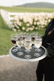A person is holding a tray with several glasses of white and red wine. The setting appears to be outdoors with a scenic background of a white fence, lush greenery, and flowers in soft focus.