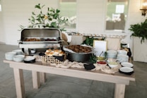 A buffet table is set up with various dishes and condiments. There are plates stacked on one end and a display of sandwiches or sliders in a basket. In the middle, there is a large pot with cooked food, possibly falafel or meatballs, and a selection of dips in small bowls, including ketchup and possibly coleslaw. Leafy greens or decorative plants are arranged around the dishes, and a stainless steel chafing dish holds hot food.