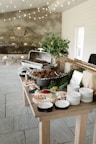 A rustic buffet setup on a wooden table with a variety of dishes. There is a chafing dish containing cooked food, surrounded by plates and bowls filled with colorful condiments like red sauce, and pickles. The decor includes leafy greens and a plant in a pot, with string lights hanging above, providing a cozy atmosphere.