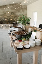 A rustic buffet setup on a wooden table with a variety of dishes. There is a chafing dish containing cooked food, surrounded by plates and bowls filled with colorful condiments like red sauce, and pickles. The decor includes leafy greens and a plant in a pot, with string lights hanging above, providing a cozy atmosphere.