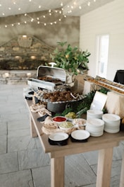 A rustic buffet setup on a wooden table with a variety of dishes. There is a chafing dish containing cooked food, surrounded by plates and bowls filled with colorful condiments like red sauce, and pickles. The decor includes leafy greens and a plant in a pot, with string lights hanging above, providing a cozy atmosphere.