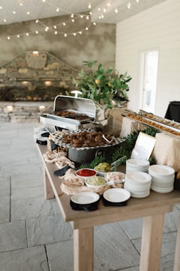 A rustic buffet setup on a wooden table with a variety of dishes. There is a chafing dish containing cooked food, surrounded by plates and bowls filled with colorful condiments like red sauce, and pickles. The decor includes leafy greens and a plant in a pot, with string lights hanging above, providing a cozy atmosphere.