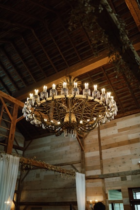 A rustic chandelier hanging in a warmly lit living room with wooden beams.