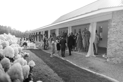 A gathering of people, some in formal attire, is taking place near a long porch with hanging plants and decorated columns. On the left, hydrangea flowers are visible, indicating a garden setting. Tables with chairs are arranged on the grass lawn, suggesting the event is a social outdoor gathering like a wedding or party.