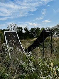 A grassy field with a metal ramp structure in the foreground. The word 'MARS' is painted on the ramp, which is surrounded by wildflowers and tall grasses. Trees and a partly cloudy sky can be seen in the background.