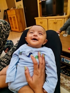 A baby wearing a light blue outfit adorned with dinosaur designs is being held by an adult with mint-green painted nails. The baby is smiling with a joyful expression. The setting appears to be a cozy living room with wooden furniture, a television, and a patterned rug. Other adults and objects like a drinking glass and books can be seen in the background.