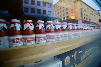 A row of glass bottles filled with tomato sauce is neatly arranged on a wooden shelf. Each bottle has a white and red label and is topped with a white paper cover secured with a red tie. The reflection of buildings outside is visible on the glass, adding an urban background to the display.