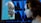 A futuristic scene where a human-like robot face emerges from a computer screen, closely facing a woman with brown hair. The background consists of a grid of blue-lit panels.