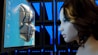 A futuristic scene where a human-like robot face emerges from a computer screen, closely facing a woman with brown hair. The background consists of a grid of blue-lit panels.