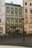 A historic European street scene featuring a row of buildings with ornate architectural details. A green building is in the center with multiple windows and signs on the ground level for shops, including 'pierre cardin.' A tall street lamp stands in the foreground, and small barriers line the edge of the pavement.