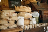 A rustic scene featuring a variety of freshly baked bread loaves stacked on a wooden table next to jars covered with lace cloth. A miniature wooden house adds a quaint element, and a chalkboard sign with menu items like onion, cucumber, sausage, and meat is visible in the background.