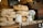A rustic scene featuring a variety of freshly baked bread loaves stacked on a wooden table next to jars covered with lace cloth. A miniature wooden house adds a quaint element, and a chalkboard sign with menu items like onion, cucumber, sausage, and meat is visible in the background.