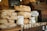 A rustic scene featuring a variety of freshly baked bread loaves stacked on a wooden table next to jars covered with lace cloth. A miniature wooden house adds a quaint element, and a chalkboard sign with menu items like onion, cucumber, sausage, and meat is visible in the background.